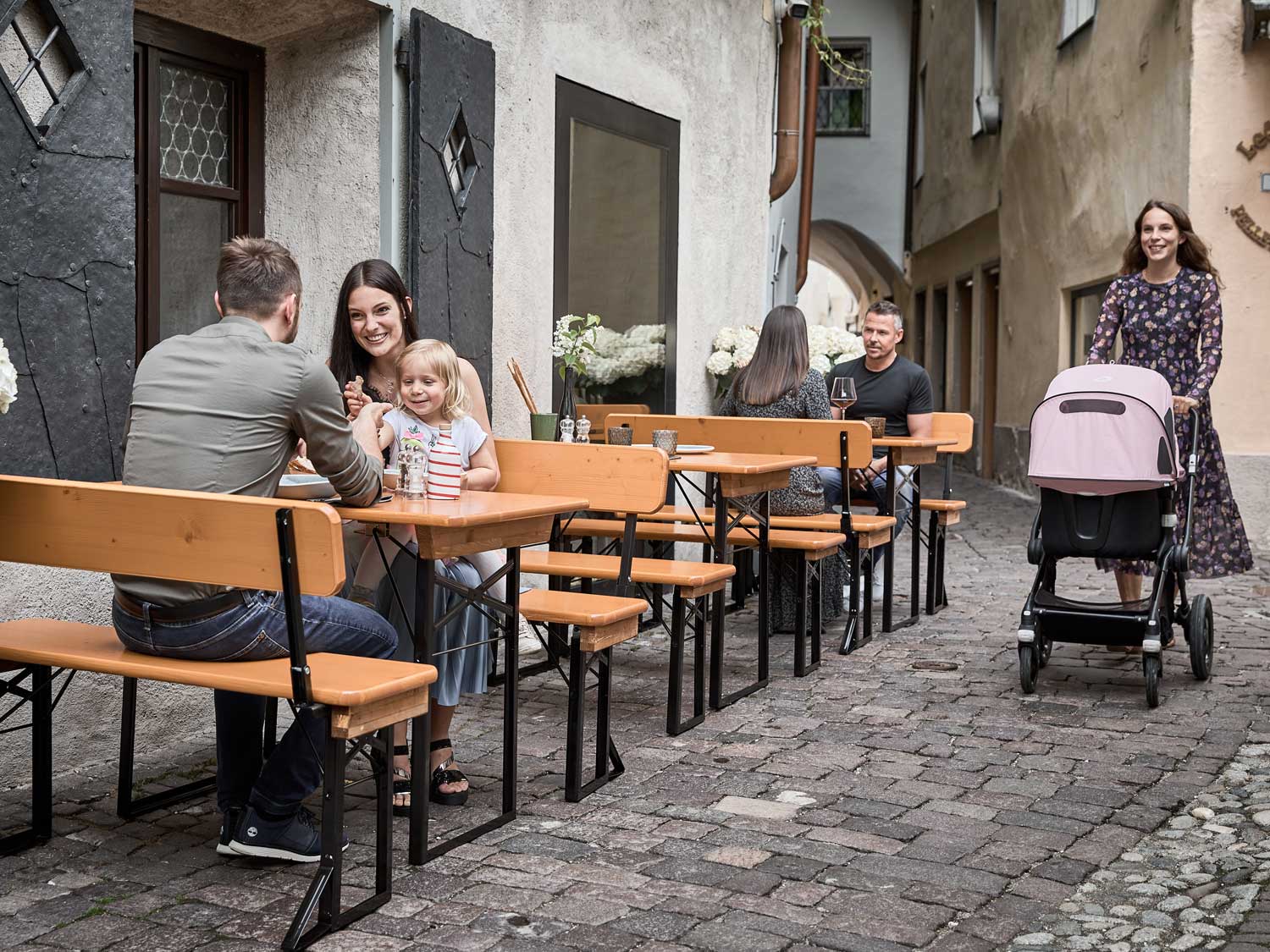 Mehrere Personen sitzen auf kleinen Bierzeltgarnituren in einer Gasse bei einem Restaurant.