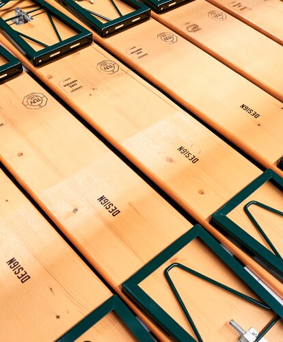 Folded classic beer benches with green frame, seats facing down, folding mechanisms visible The photo shows a row of folded beer benches with green frames and seat surfaces facing downward.