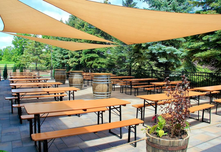 L'area esterna di Elm Creek Brewing presenta diversi set da birreria Classic in colore pino con telai neri, disposti sotto grandi vele ombreggianti. L'area è incorniciata da alberi verdi.