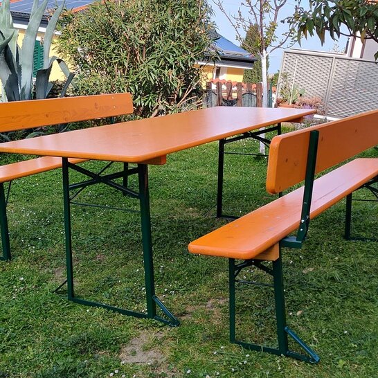 Eine breite Bierzeltgarnitur steht in einem Garten Eine breite Bierzeltgarnitur in der Farbe Ocker mit Rückenlehne und grünem Untergestell.